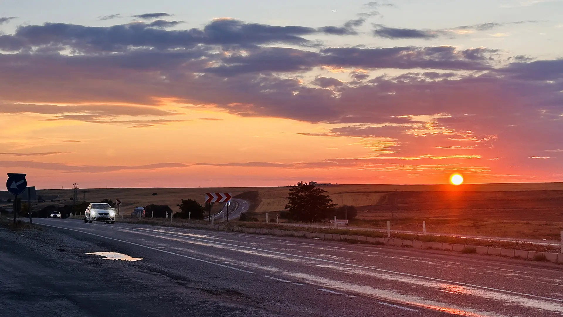 Atemberaubender Sonnenuntergang über einer Landstraße. In der Ferne ist ein Auto zu sehen, das auf der kurvenreichen Straße fährt.